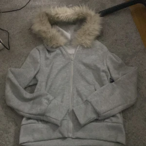 Fur zip up grå - Mysig grå hoodie med dragkedja och en fluffig pälsfodrad huva. Perfekt för kyliga dagar när du vill hålla dig varm och bekväm. Den har långa ärmar och en avslappnad passform.