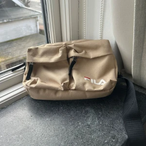 Beige axelväska/magväska från Fila - Säljer en beige axelväska/magväska från Fila med justerbar svart rem och praktiska dragkedjor. Perfekt för att bära med sig det nödvändigaste. Väskan har en stilren design med Fila-loggan på framsidan.