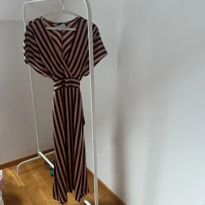 Randig långklänning från Neo Noir - Snygg långklänning från Neo Noir med ett randigt mönster i vinrött och beige. Klänningen har en v-ringad design och korta ärmar, perfekt för en stilren look.