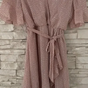 Rosa klänning med knytband - Söt rosa klänning med småmönstrat tryck och volangdetaljer. Den har en v-ringad hals och korta ärmar. Knytband i midjan för en smickrande passform.