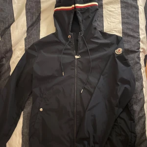 Svart vindjacka från Moncler - Snygg mörk blå vindjacka från Moncler med dragkedja och huva. Jackan har Moncler-logga på ärmen och en justerbar dragsko i huvan. Perfekt för blåsiga dagar. Jackan är såklart äkta, skriv till mig för mer information.