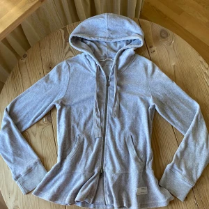 Grå peplum hoodie från Odd Molly - Säljer en grå hoodie från Odd Molly med dragkedja och huva. Tröjan har en söt rosett på baksidan och ribbade muddar vid ärmsluten. Perfekt för en avslappnad stil. 🩶