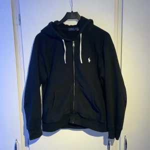 Svart hoodie från Ralph Lauren - Säljer en stilren svart hoodie från Ralph Lauren med dragkedja och vit logga på bröstet. Den har en justerbar huva med vita snören och praktiska fickor framtill. Perfekt för en avslappnad look. Pris kan diskuteras vid snabb affär✌🏽