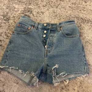 Blå jeansshorts från Levi's - Säljer ett par snygga blå jeansshorts från Levi's med hög midja och fransiga kanter. Shortsen har knappar framtill och klassiska bakfickor med Levi's logga. Perfekta för en avslappnad stil.