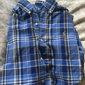 Blå rutig skjorta från Ralph Lauren - Säljer en snygg blå rutig skjorta från Ralph Lauren. Skjortan har långa ärmar och klassiska knappar framtill. Den är broderad med den ikoniska loggan på bröstet. Perfekt för en stilren look! ALDRIG ANVÄND!