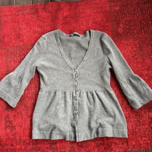 Grå babydoll kofta med knappar - Säljer en stilren grå babydoll kofta med v-ringning och knappar framtill. Koftan har trekvartsärmar och en lätt utsvängd form nedtill. Perfekt för lager-på-lager under kyligare dagar.