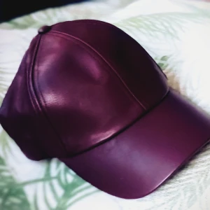 Mörk lila keps i syntet skinn - Snygg mörk lila keps i syntetiskt skinnmaterial med en klassisk design. Perfekt för att ge din outfit en stilren touch. Kepsen har en rundad skärm och justerbar passform.