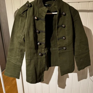 Olivgrön jacka från Gina Tricot - Snygg olivgrön jacka från Gina Tricot med dekorativa knappar framtill. Jackan har en militärinspirerad design med axelklaffar och långa ärmar. Perfekt för en stilren look.