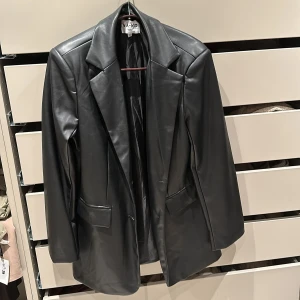 Svart skinnjacka från NA-KD - Snygg svart skinnjacka från NA-KD med klassisk kavajdesign. Jackan har långa ärmar och en stilren look som passar perfekt för en trendig outfit. Perfekt för dig som vill ha en edgy touch i garderoben.