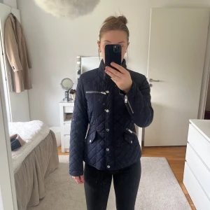 Mörkblå quiltad jacka med dragkedjor - Snygg mörkblå quiltad jacka med silverfärgade dragkedjor och knappar. Jackan har en hög krage och flera praktiska fickor. Perfekt för kyligare dagar.
