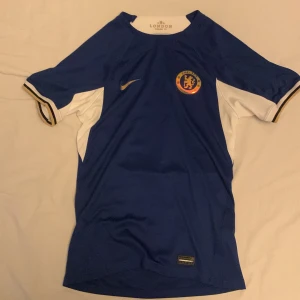 Blå Chelsea FC fotbollströja från Nike - Säljer en blå Chelsea FC fotbollströja från Nike med korta ärmar och vitt inslag vid ärmarna. Tröjan har klubbens emblem på bröstet och är tillverkad med Dri-FIT-teknologi för optimal komfort. Perfekt för alla Chelsea-fans! 💙