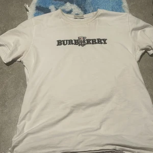 Vit t-shirt från Burberry - Säljer en stilren vit t-shirt från Burberry med deras ikoniska logga tryckt i svart på bröstet. Perfekt för en casual look. Passar bra till jeans eller shorts.