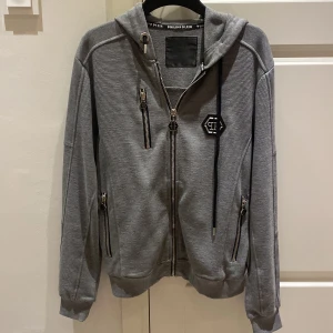 Grå huvtröja från Philipp Plein - Säljer en grå huvtröja från Philipp Plein med dragkedja och flera fickor. Tröjan har en broderad logga på bröstet och ribbade muddar. 