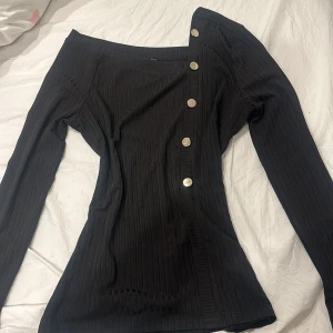 Svart ribbad offshoulder topp - Snygg svart ribbad offshoulder topp med knappar längs ena sidan. Den har långa ärmar och en stilren design som passar perfekt för en trendig look.