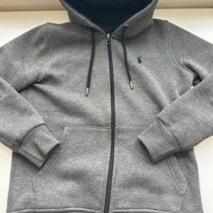 Grå hoodie från Ralph Lauren - Säljer en stilren grå hoodie från Ralph Lauren med dragkedja och justerbar huva. Tröjan har ribbade muddar och en liten broderad logga på bröstet. Perfekt för en avslappnad look.