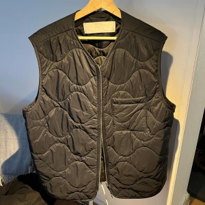 Svart quiltad väst ARKET - Snygg svart quiltad väst med dragkedja framtill. Västen har ett vågigt mönster och en praktisk bröstficka. Perfekt för lager-på-lager under kyligare dagar.