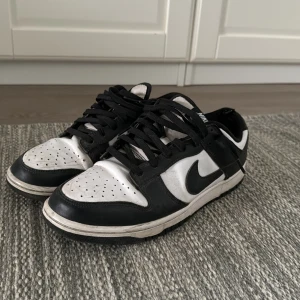 Svartvita Nike Dunk sneakers - Snygga svartvita Nike Dunk sneakers med klassisk design. Skorna har en vit bas med svarta detaljer och snörning. Perfekta för en stilren look. Skorna är inte tvättade än och har slitningar men säljer de nu för ett schysst pris! Hör av er vid intresse! 