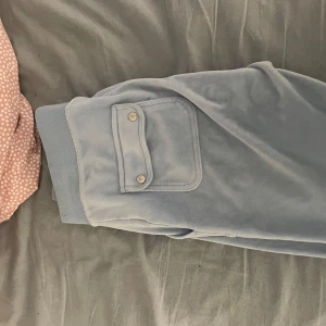 Blå juicy med fickdetalj - Säljer ett par bekväma blå juicybyxor med en praktisk ficka på sidan. Byxorna har en avslappnad passform och är perfekta för en mysig dag hemma. Fickan har två knappar för extra säkerhet. Storlek XS passar även S