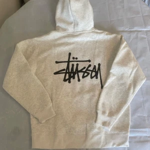 Grå hoodie från Stussy - Säljer en snygg grå hoodie från Stussy med deras ikoniska logga tryckt på bröstet och ryggen. Hoodien har en klassisk känguruficka och justerbar huva med dragsko. Perfekt för en avslappnad stil.