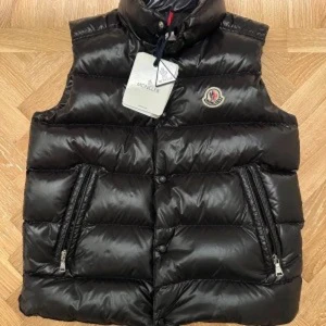 Svart dunväst från Moncler - Snygg svart dunväst från Moncler med hög krage och dragkedja. Västen har två praktiska fickor med dragkedjor och en broderad logotyp på bröstet. Perfekt för kyligare dagar.