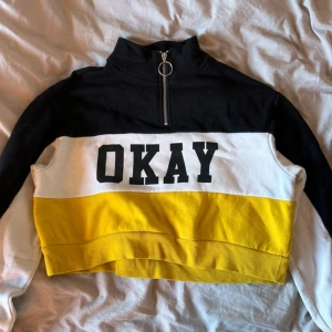 Svart, vit och gul croppad tröja med text croptop - Säljer en cool croppad tröja i svart, vit och gul med texten 'OKAY' på bröstet. Tröjan har en dragkedja framtill och långa ärmar. Perfekt för en avslappnad stil!