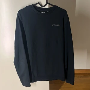 Mörk blå tröja från Lyle & Scott - Säljer en stilren mörkblå tröja från Lyle & Scott. Tröjan har en diskret logga på bröstet och en liten gul detalj på ärmen. Perfekt för en avslappnad look. Köpt för 500 kr 