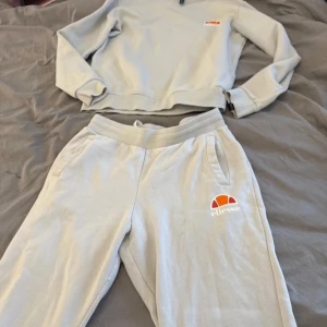 Tvådelat set från Ellesse - Säljer en stilren grå sweatshirt från Ellesse med deras ikoniska logga i vitt och orange på bröstet. Tröjan har långa ärmar och en rund halsringning. Perfekt för en avslappnad look. Och byxan