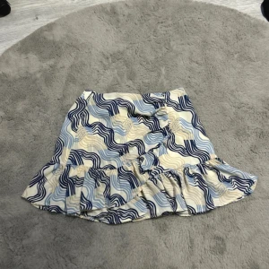 Mönstrad kjol med volang - Snygg kortkjol med vågigt mönster i blått och beige. Kjolen har volangdetaljer och knappar framtill som ger en unik look. Perfekt för att liva upp garderoben!