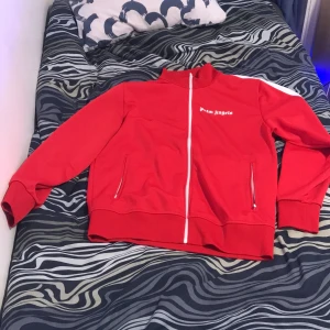 Röd track jacket från Palm Angels - Snygg röd track jacket från Palm Angels med vit dragkedja och vita detaljer på ärmarna. Jackan har två fickor med dragkedjor och en hög krage. Perfekt för en sportig look.
