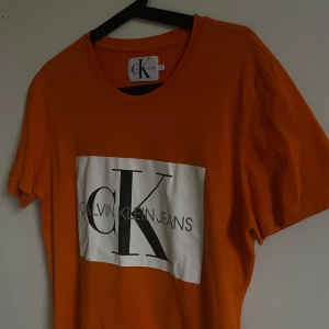 Orange t-shirt från Calvin Klein Jeans - Snygg orange t-shirt från Calvin Klein Jeans med stort tryck på framsidan. Perfekt för en avslappnad stil. Kortärmad och tillverkad i mjukt material.