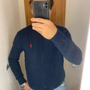 Mörkblå stickad tröja från Ralph Lauren - Säljer en snygg mörkblå stickad tröja från Ralph Lauren med det klassiska röda logotypen på bröstet. Tröjan har en kabelstickad design och rund halsringning. Perfekt för kyligare dagar! 
