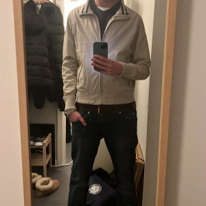 Beige bomberjacka med dragkedja - Säljer en stilren beige bomberjacka med dragkedja och ribbade muddar. Jackan har en broderad detalj på ärmen och en klassisk krage. Perfekt för en avslappnad look.