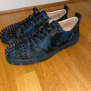 Camo Christian Louboutin - Snygga sneakers i camouflage från Christian Louboutin med nitar på tån och klassisk röd sula. Skorna har snörning och kommer med originalförpackning, kvitto och dustbag. Andvänd 1 gång.