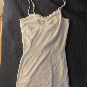 Vit kortklänning  - Säljer en söt vit klänning med smala axelband och ett diskret sött mönster. Klänningen har en åtsittande passform och är mycket kort och söta detaljer vid bröstet. 