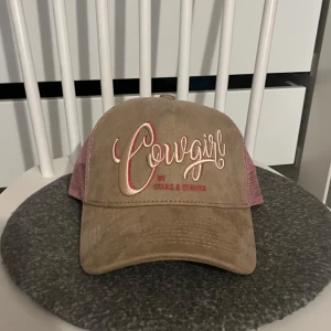 Beige och rosa keps med text - Snygg keps i beige och rosa med texten 'Cowgirl' broderad framtill. Märket är Stars & Stripes. Kepsen har ett nätmaterial baktill för extra ventilation.
