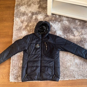 Svart pufferjacka från Sail Racing - Snygg svart pufferjacka från Sail Racing med huva och dragkedja. Jackan har en quiltad design och är perfekt för kyligare väder. Den har praktiska fickor med dragkedjor och en stilren logga på bröstet.