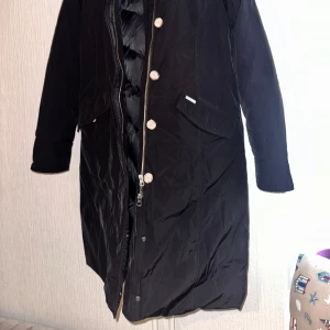 Svart parkajacka med pälsdetalj - Säljer en stilren svart parkajacka från woolrich. Jackan har en mysig pälsdetalj på huvan och stängs med både dragkedja och knappar. Perfekt för kyliga dagar!