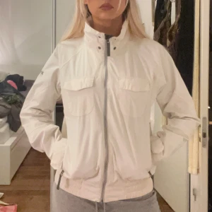 Vit vindjacka med dragkedja - Snygg vit vindjacka med dragkedja och hög krage. Jackan har praktiska fickor med lock och ribbade muddar vid ärmslut och nederkant. Perfekt för blåsiga dagar.