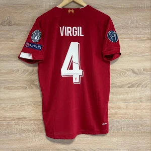 Liverpool Hemma  Virgil 4 XL - Säljer en röd Liverpool fotbollströja från New Balance med Virgil och nummer 4 på ryggen. Tröjan har korta ärmar och Champions League-märken på ärmarna. Framsidan pryds av Standard Chartered-loggan och klubbmärket.