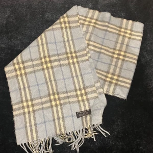 Rutig halsduk från Burberry - Snygg rutig halsduk från Burberry i ljusblått, grått och beige. Den har klassiska fransar i ändarna och är perfekt för att hålla dig varm under kyliga dagar. Ett stilrent val för den modemedvetna.