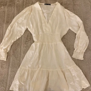 Vit linne-klänning från Zara - Elegant vit klänning från Zara gjord av linne, med v-ringning och långa ärmar. Klänningen har en smockdetalj i midjan och volang nedtill, vilket ger en fin siluett. Perfekt för en stilren look. Säljer då den tyvärr blivit för liten 💗