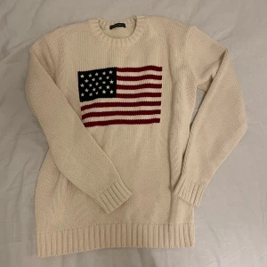 Vit stickad tröja från Brandy Melville - Säljer en vit stickad tröja från Brandy Melville med amerikanska flaggan på framsidan. Tröjan har långa ärmar och ribbstickade detaljer vid hals, ärmslut och nederkant. Använd en gång, tröjan är alltså i nyskick. Säljer pga den är för stor för mig.
