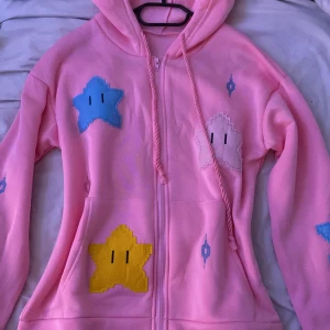 Rosa hoodie med stjärnmönster - Säljer en rosa hoodie med dragkedja och färgglada stjärnor. Aldrig haft på mig och har bara köpt den någon månad sen så jag skulle säga att den är nytt skick, priset kan diskuteras