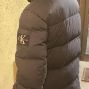 Svart dunjacka från Calvin Klein - Snygg och varm svart dunjacka från Calvin Klein Jeans. Jackan har en klassisk pufferdesign med långärmade ärmar och en diskret logga på ärmen. Perfekt för kyliga dagar.