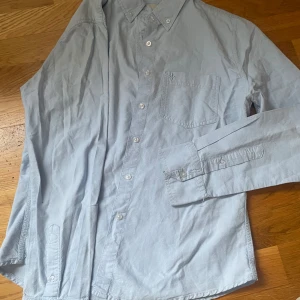 BUDA PRIS: Ljusblå skjorta med bröstficka - Snygg ljusblå skjorta med klassisk krage och knappar framtill. Skjortan har en praktisk bröstficka med en diskret broderad detalj. Perfekt för en stilren look.