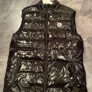 Svart dunväst från Moncler - Snygg svart dunväst från Moncler med glansig yta och dragkedja framtill. Västen har ett broderat Moncler-märke på bröstet och praktiska fickor med dragkedja. Perfekt för kyligare dagar. Västen har en ytterst liten defekt som ni kan se ovanför den högra fickan. Priset kan diskuteras. Hör gärna av er vid frågor😊