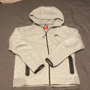 Nike tech - Säljer en grå huvtröja från Nike med dragkedja och svart logga på bröstet. Tröjan har långa ärmar och två sidofickor med dragkedja. Perfekt för en avslappnad stil.