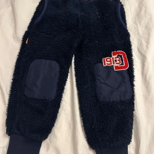 Mörkblå fleecebyxor från Didriksons - Mysiga mörkblå fleecebyxor från Didriksons med förstärkta knän och en cool broderad detalj med siffrorna '93' i rött och vitt. Perfekta för kalla dagar när du vill hålla dig varm och bekväm. De kommer tvättas och strykas innan de skickas! ⭐️