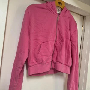 Rosa hoodie med dragkedja - Säljer en rosa hoodie med dragkedja och fickor framtill. Den har en bekväm passform och är perfekt för en avslappnad stil. Den är kortare i modellen. Bra skick.