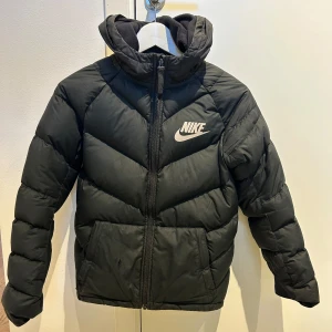 Svart dunjacka från Nike storlek 137-147 cm - Säljer en svart varm dunjacka från Nike med huva och dragkedja. Jackan har ett klassiskt Nike-märke på bröstet och är perfekt för kyligare väder. Insidan är mönstrad med Nike-loggor. Det finns lite slitage på framsidan vid fickan (se bild 4) plus 3 st väldigt små hål (se bild 5 - största hålet) Dessa går lätt att laga med lagningslapp. Jackan är junior storlek Medium 137-147 cm. Kan skicka fler bilder. 